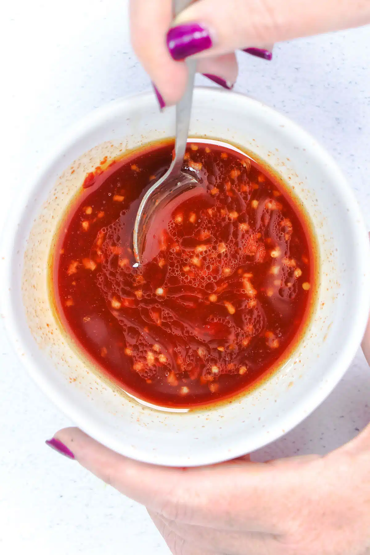 A white hand is stirring the bright red bibimbap sauce in a small white bowl.