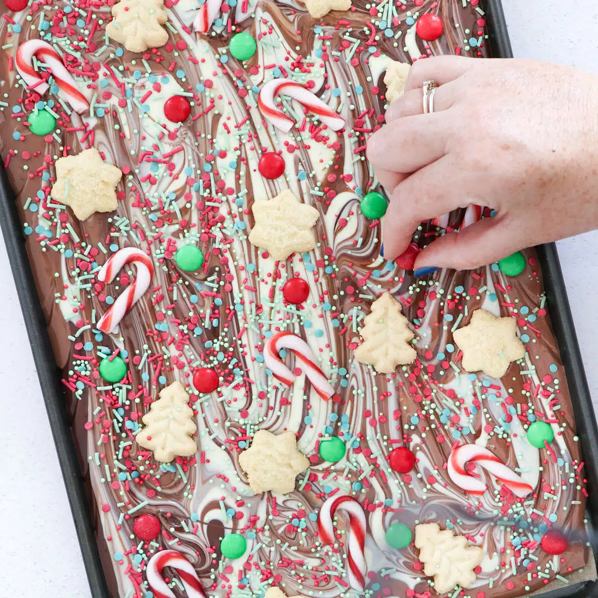 A white hand is decorating swirled chocolate bark with shortbread, candy canes, sprinkles and chocolate buds.