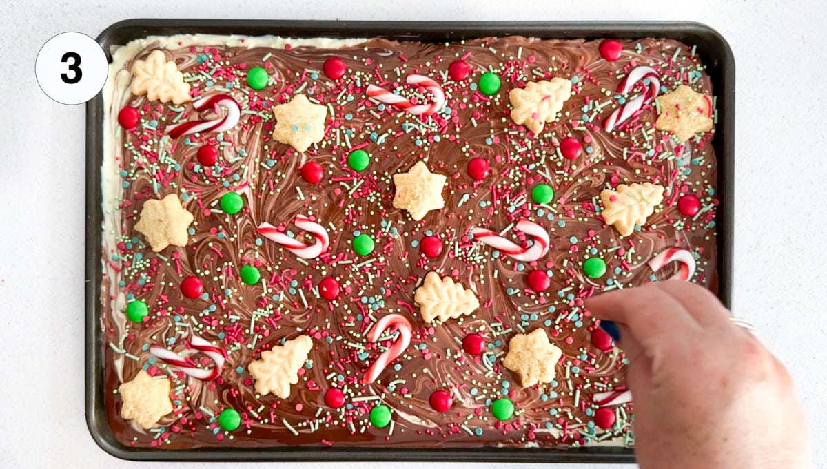 A large baking sheet is filled with decorated chocolate bark. A white hand is adding sprinkles over the top.