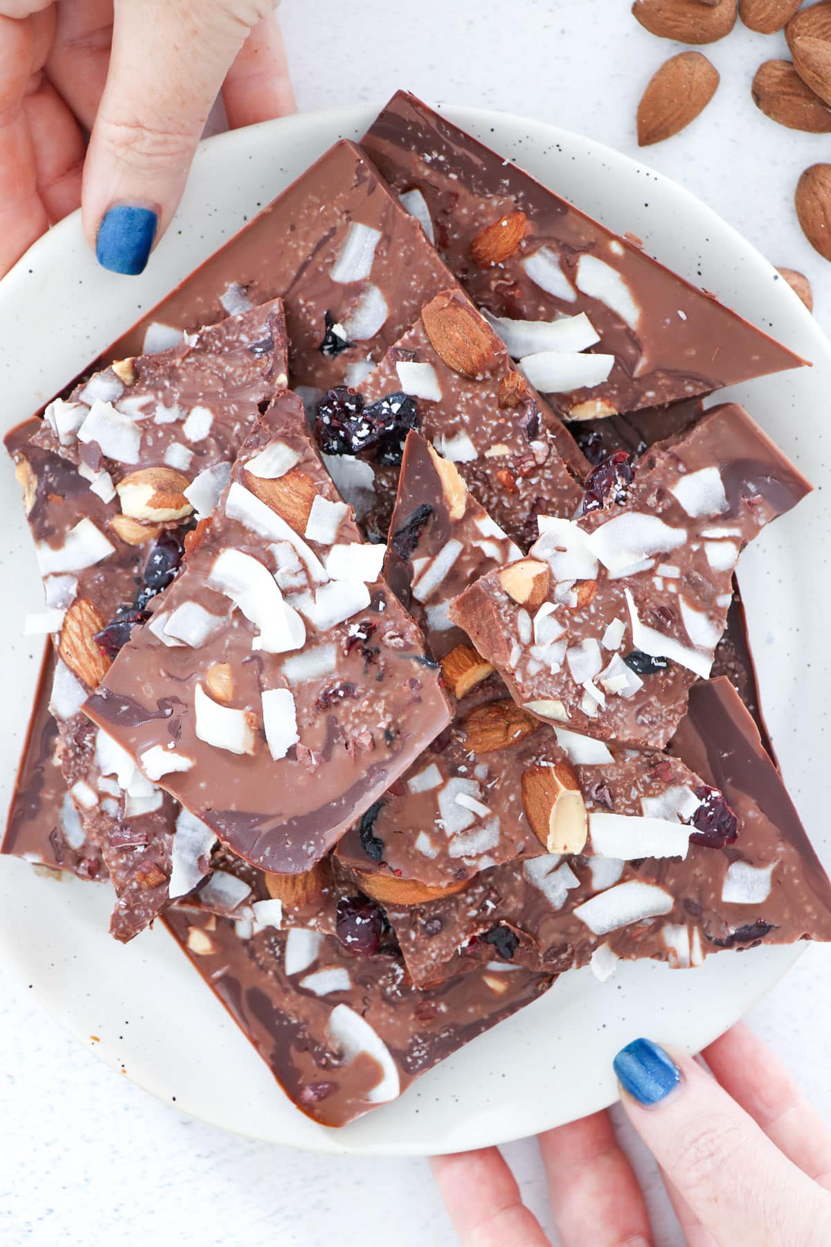 Chocolate bark topped with nuts, dried cranberries and coconut is piled on a white plate held by two white hands.