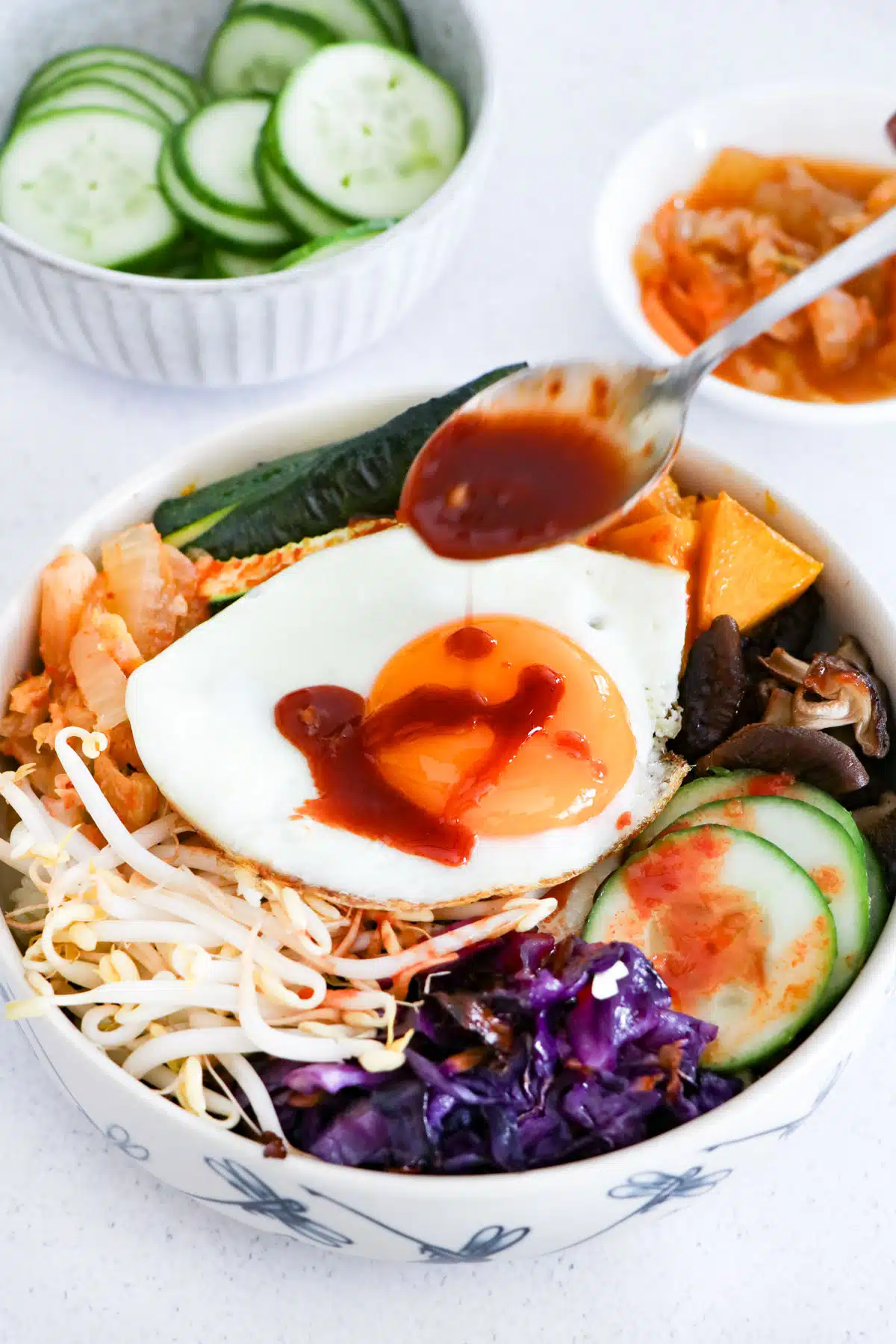 Bibimbap sauce is being drizzled over an assembled bowl of bibimbap. Bowls of pickled cucumber and kimchi can also be seen.