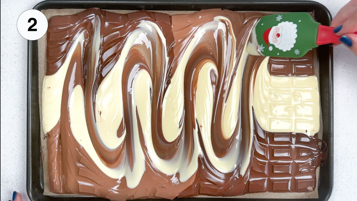 A white hand holding a spatula is swirling through milk, dark, and white chocolate laid out on a baking tray.