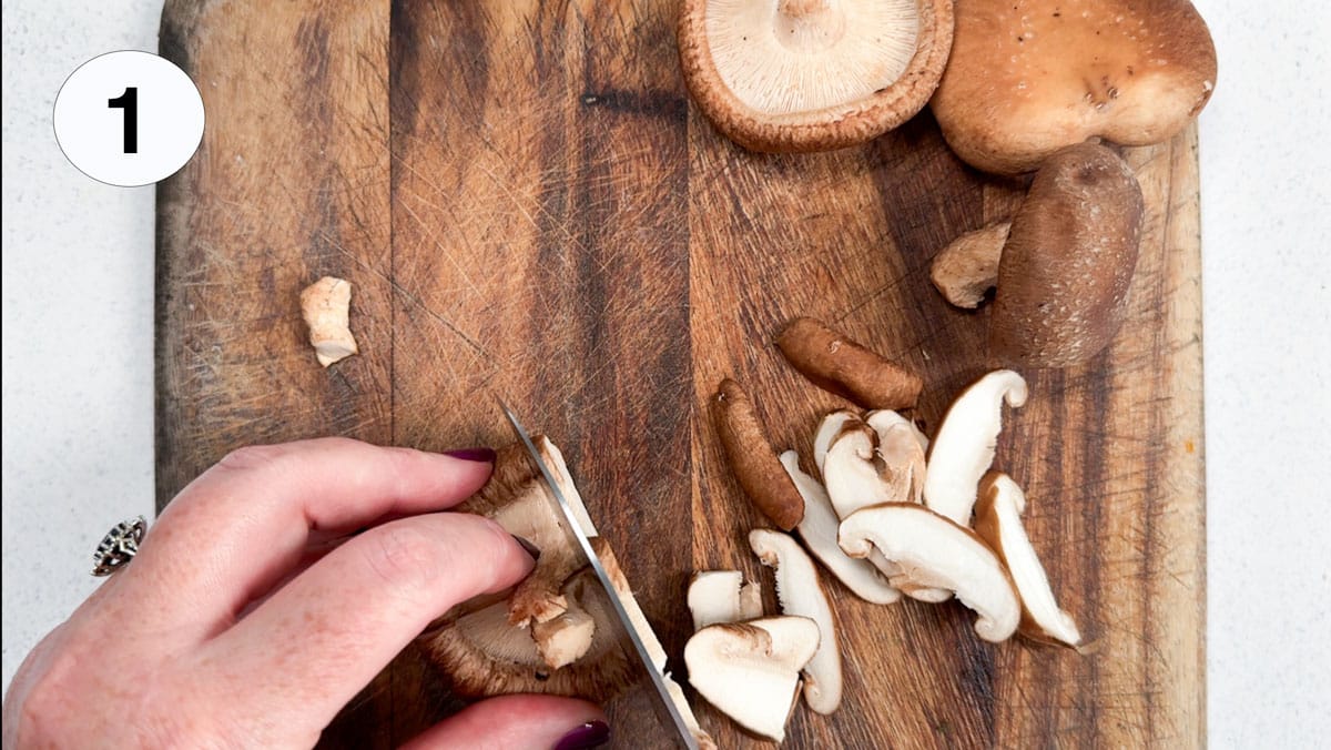 Two white hands are chopping shiitake mushrooms on a wooden chopping board.