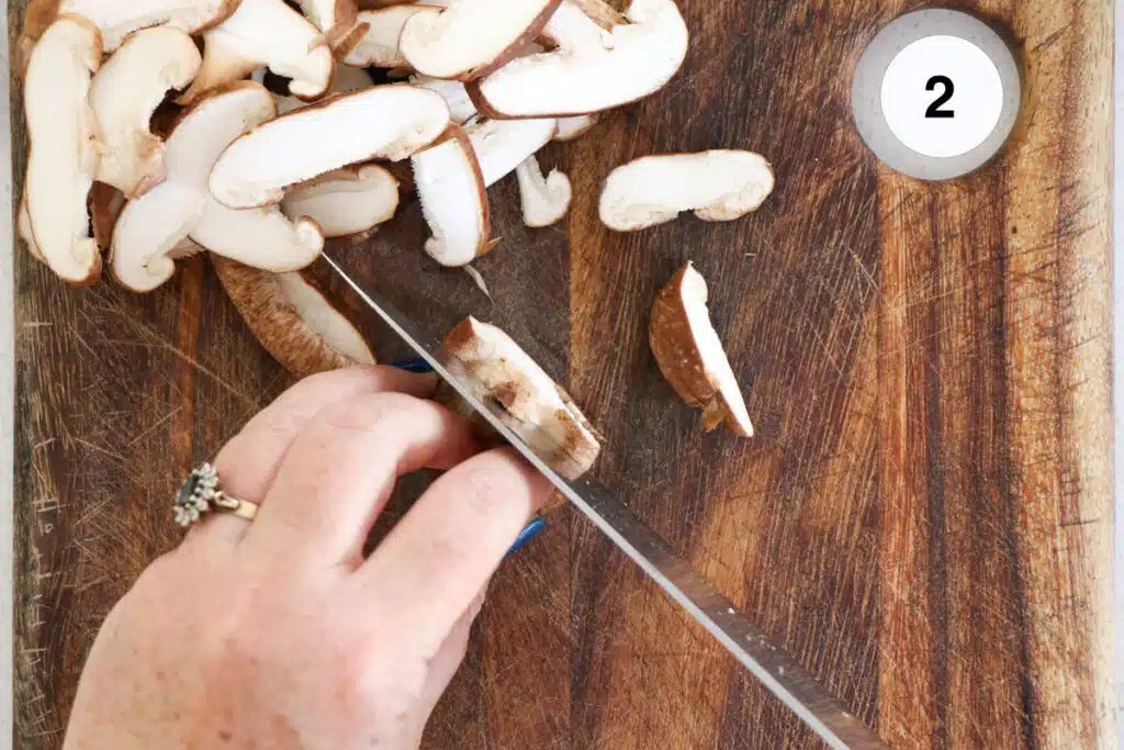 A white hand is holding a large silver knife and chopping mushrooms on a wooden chopping board.