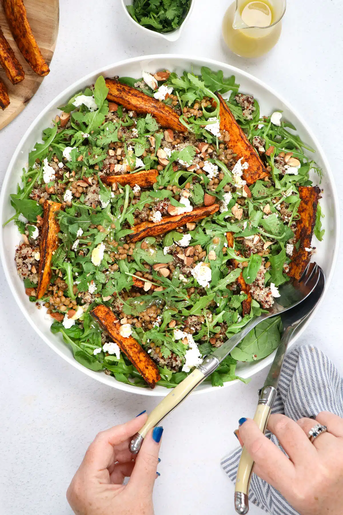 Two white hands holding salad servers are taking sweet potato quinoa salad from a large bowl.