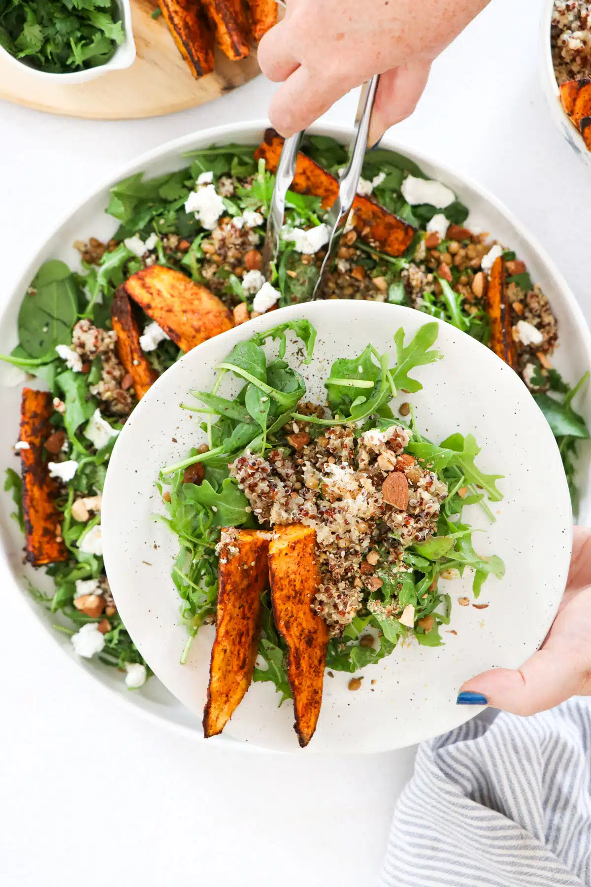 Sweet potato and quinoa salad is being served from a large bowl onto a white plate with tongs.