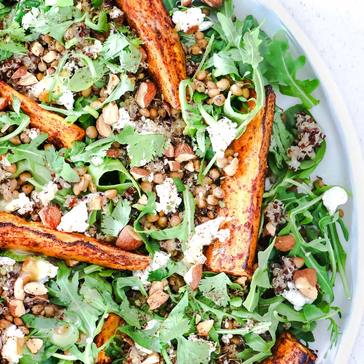 A close up looking into a bowl of sweet potato salad showing greens, potato, quinoa, lentils, goat's cheese and nuts.