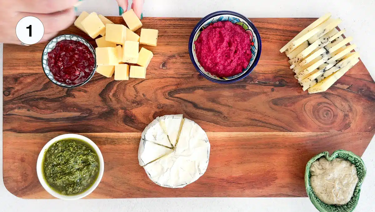 Two white hands are added cubed cheese to a large board that has bowls of dips scattered around.