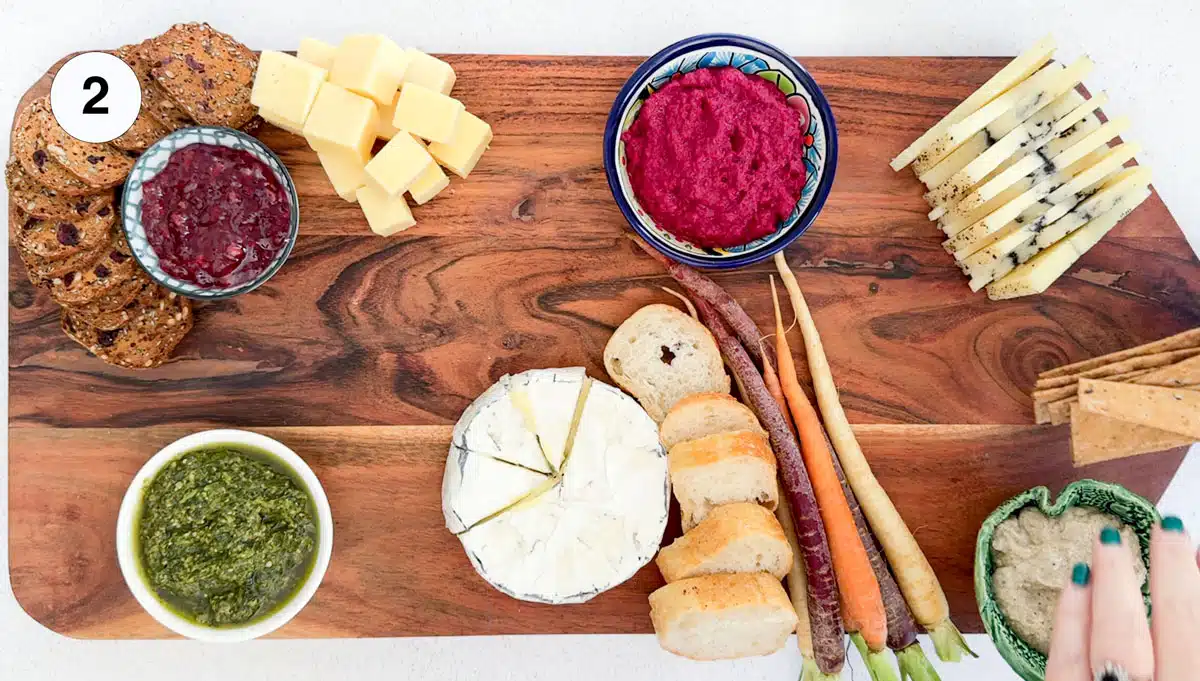 A white hand is adding crackers to a large wooden board that has dips and some veg on it.