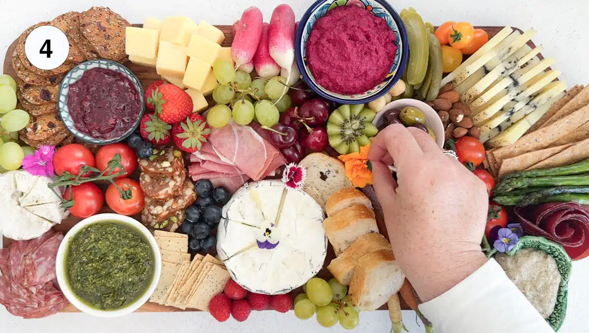 A white hand is adding edible flowers to a completed charcuterie board that's filled with a variety of ingredients.