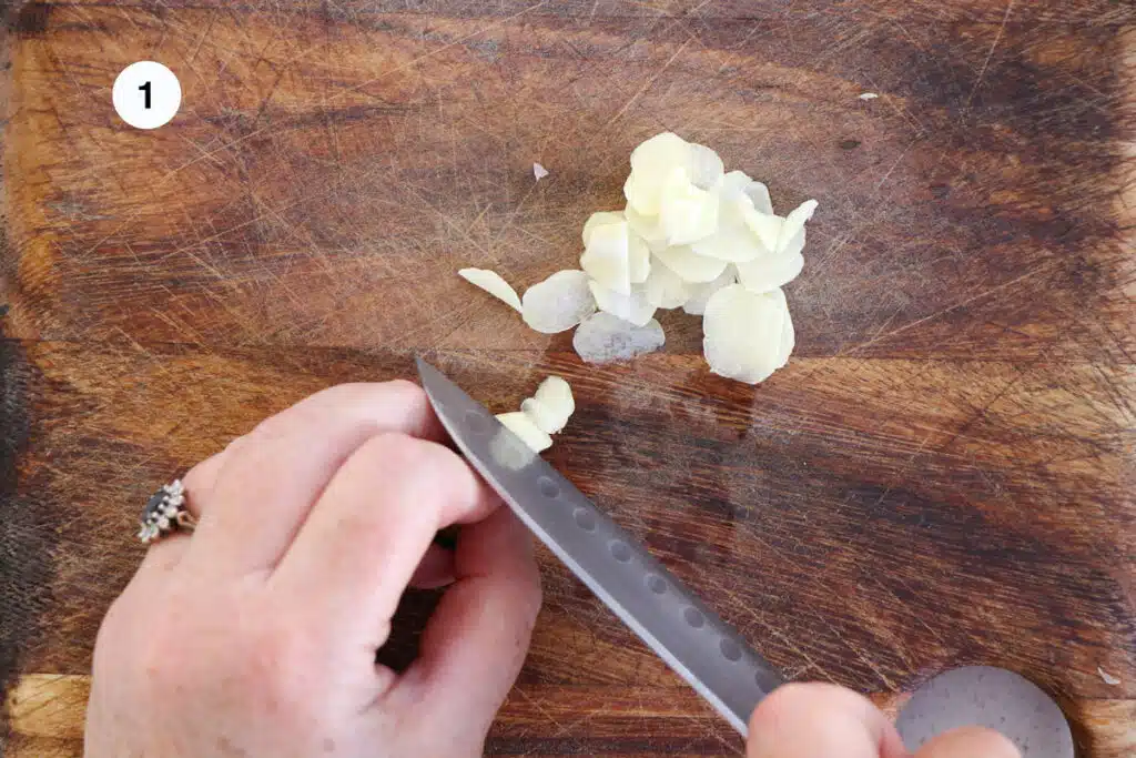 Two white hands are slicing garlic on a wooden chopping board.