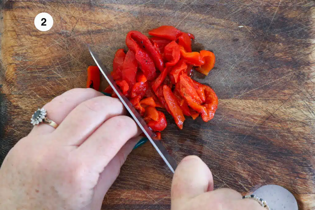 Two white hands are chopping roasted red peppers on a wooden chopping board.