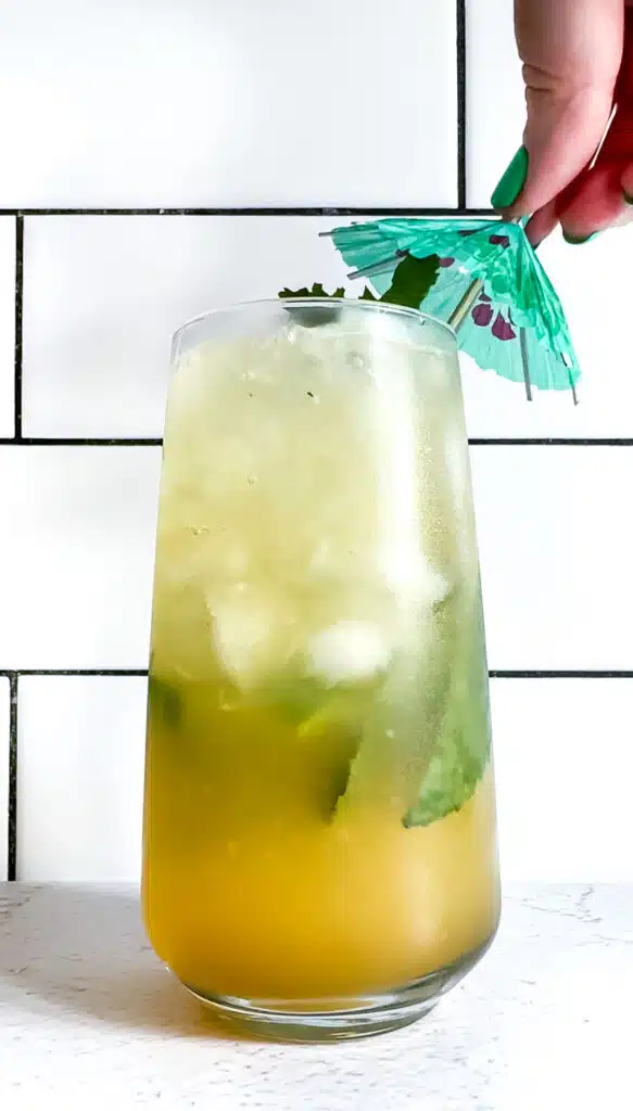 A white hand is placing a green cocktail umbrella on top of a glass filled with a ginger pineapple mocktail.