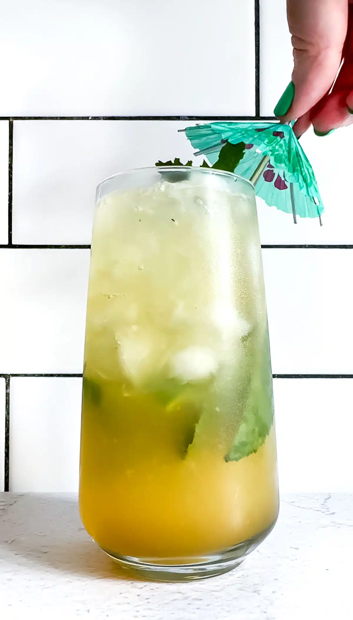 A white hand is placing a green cocktail umbrella on top of a glass filled with a ginger pineapple mocktail.