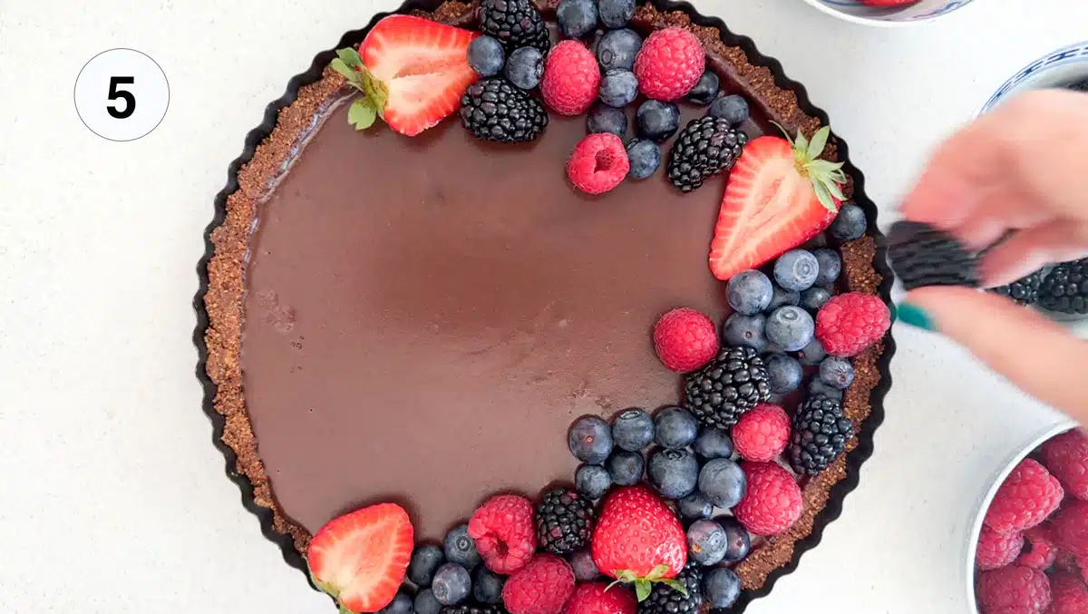 A white hand is decorating the top of the raw chocolate tart with mixed berries.