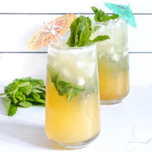 Two tall glasses sit on a white bench filled with a ginger pineapple mocktail and decorated with cocktail umbrellas.