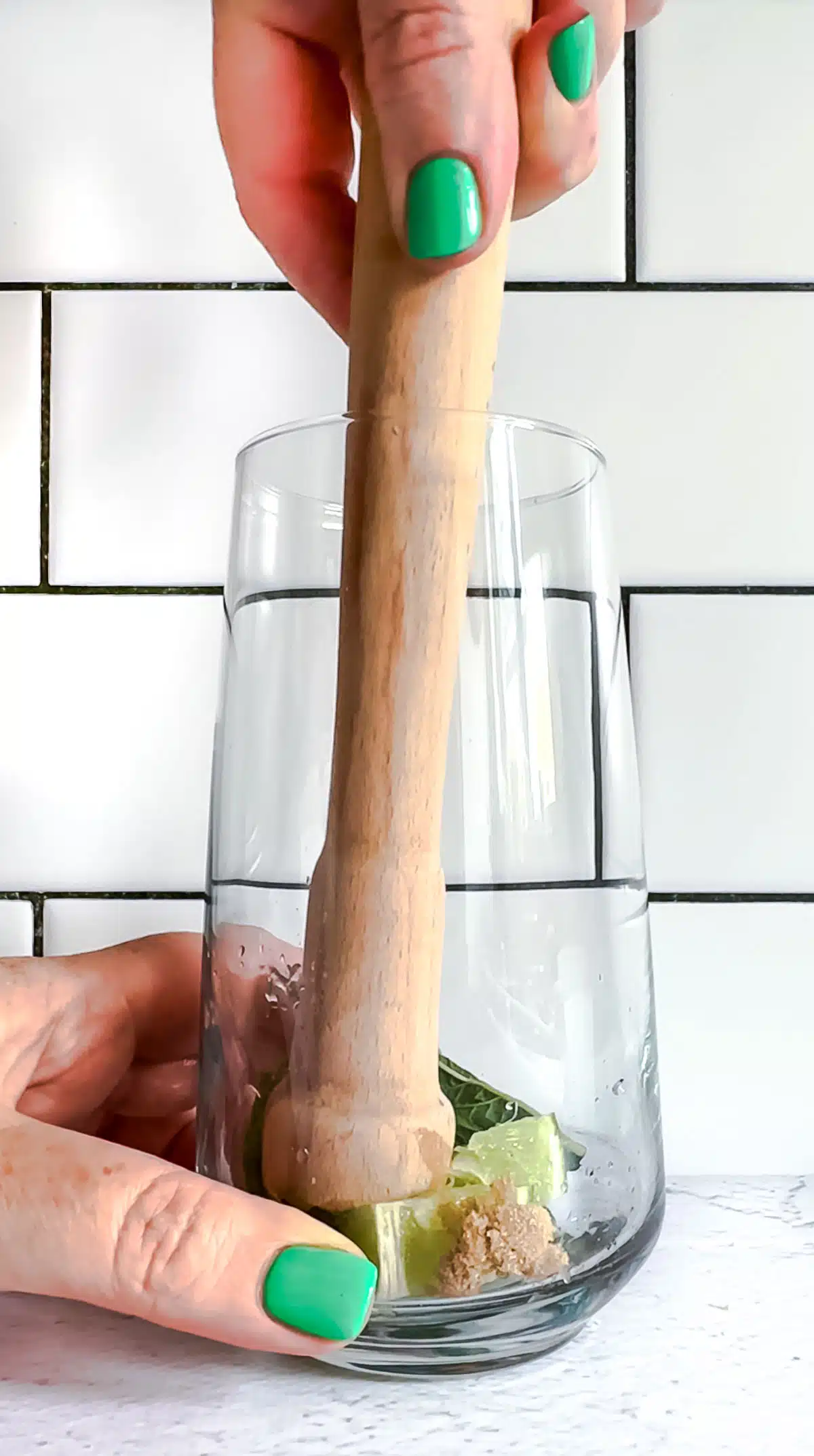 A white hand is holding a tall glass. Inside a muddle is crushing mint, limes, and brown sugar.