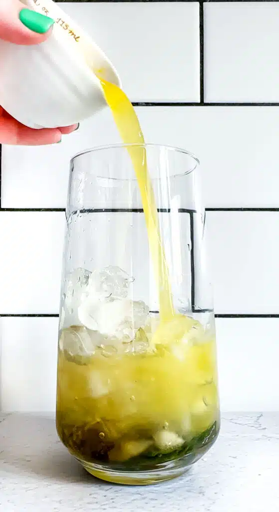 Pineapple juice is being poured from a small white measuring cup into a tall glass filled with ice and mint.