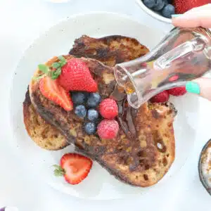 Maple syrup is being drizzled over two slices of french toast topped with mixed berries.