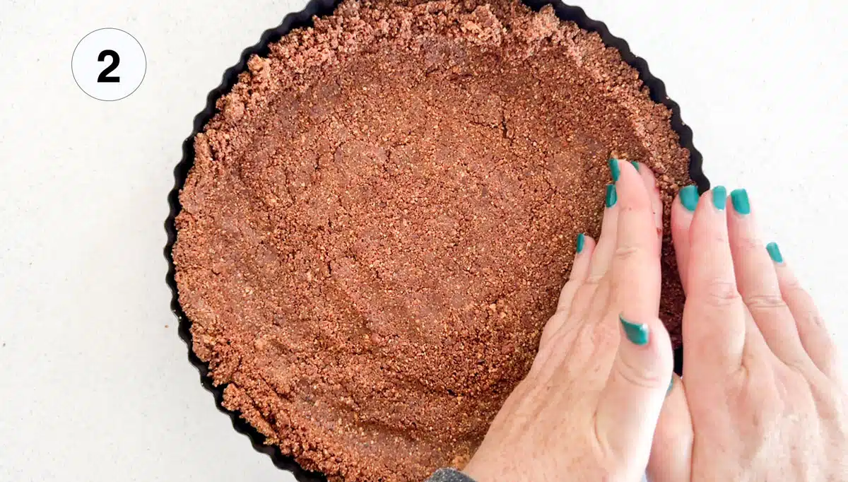 Two white hands are pressing a raw chocolate tart base into a tart tin.
