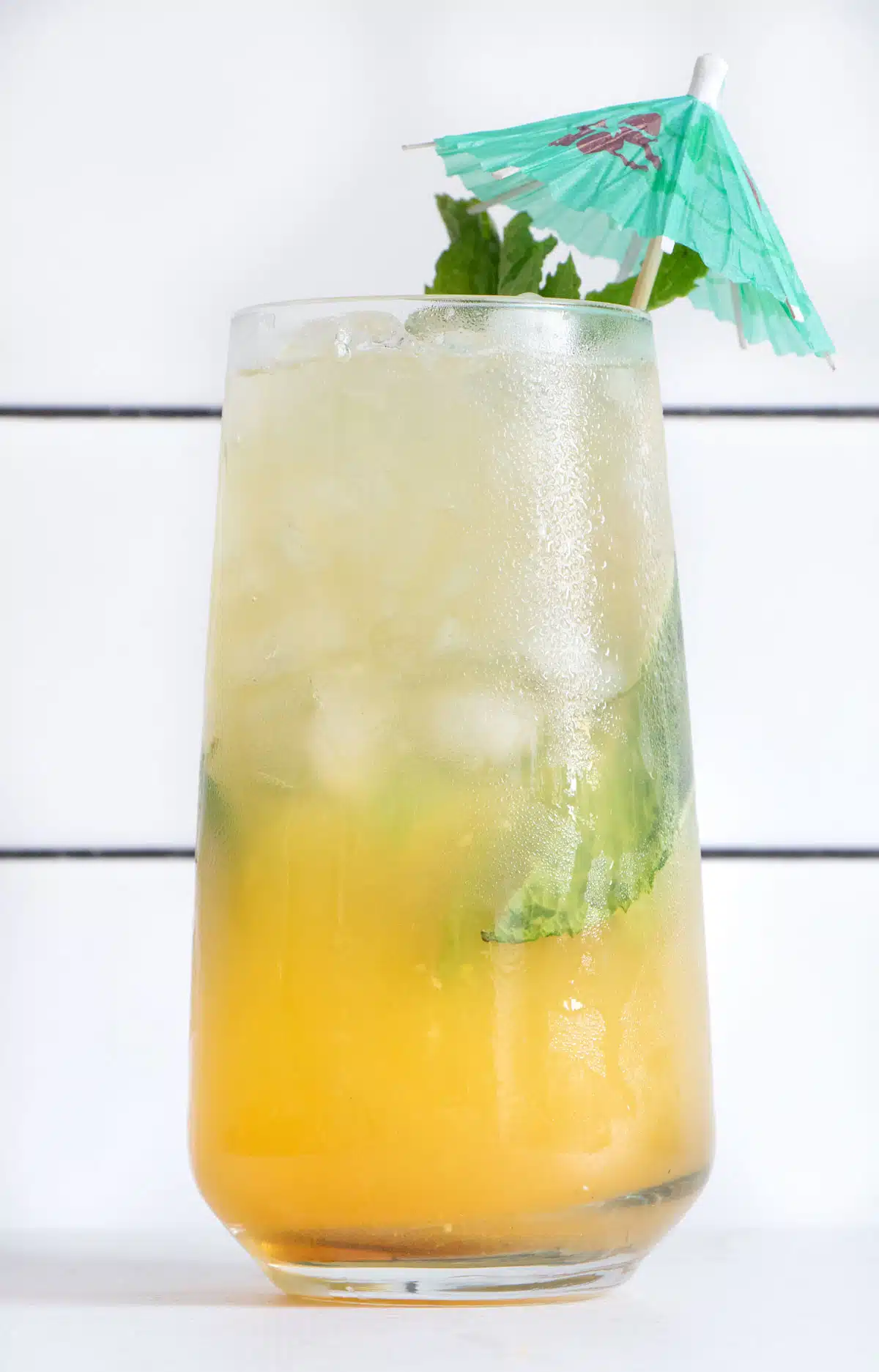A tall glass is in front of white subway tiles filled with a pineapple ginger mocktail and a green cocktail umbrella.