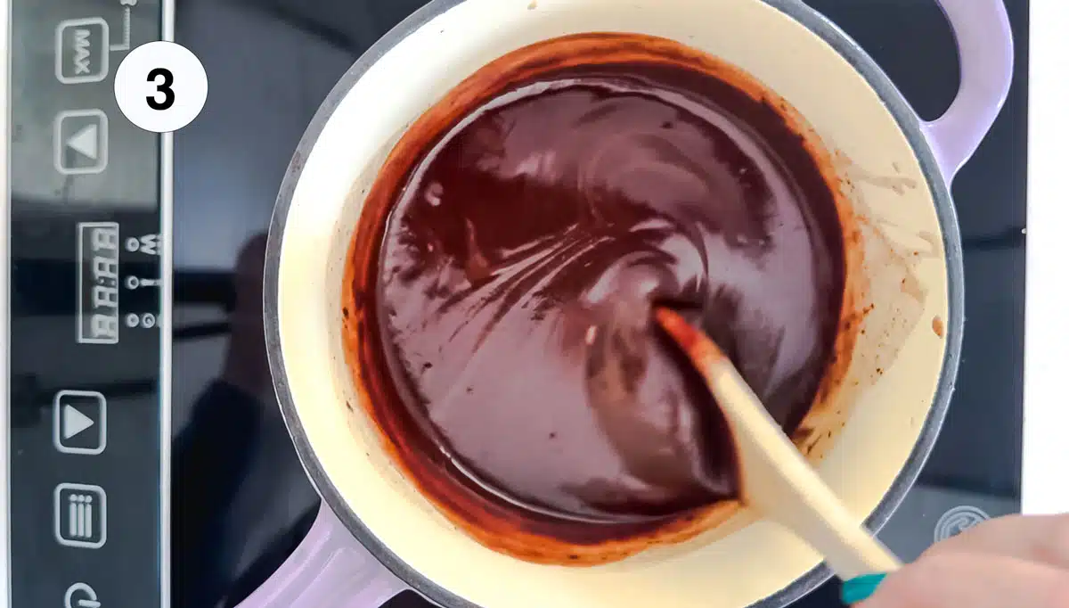 Looking down into a pot, a hand holding a wooden spoon is stirring chocolate ganache.
