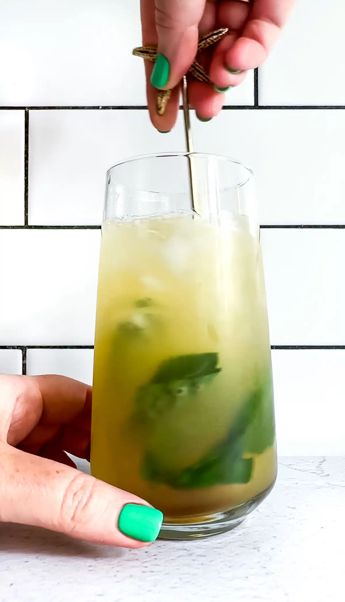A white hand holds a tall glass filled with a ginger mocktail while the other hand agitates it with a long gold stirrer.