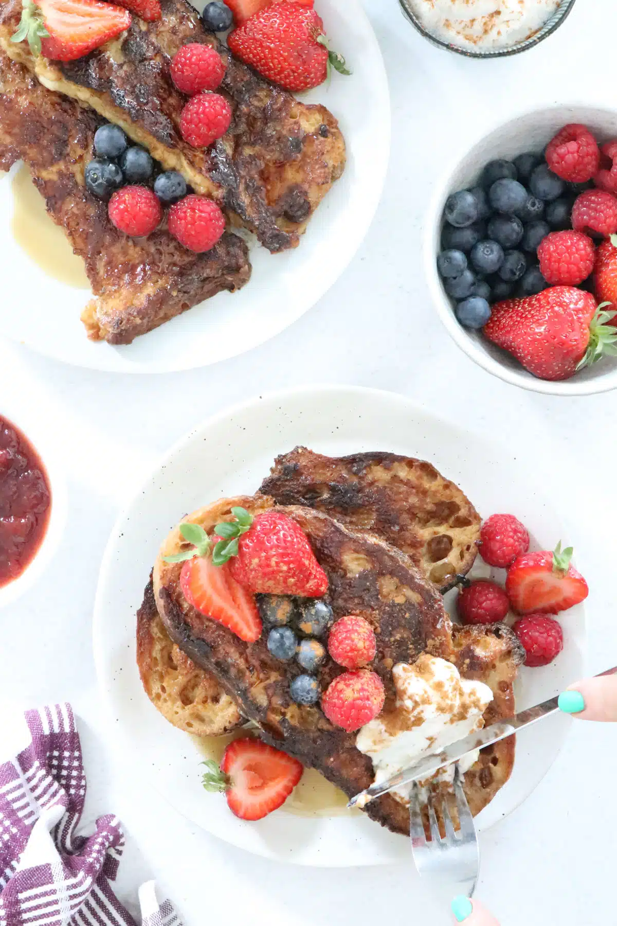 Two plates filled with sourdough french toast, berries, and coconut yoghurt are on a table surrounded by berries and jam.