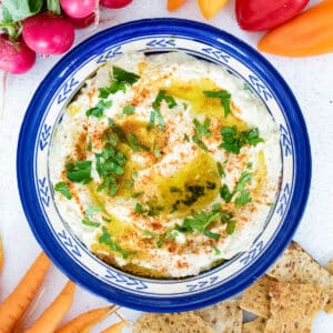 Close-up of a blue plate filled with eggplant dip. It's surrounded by crackers and colourful vegetables.
