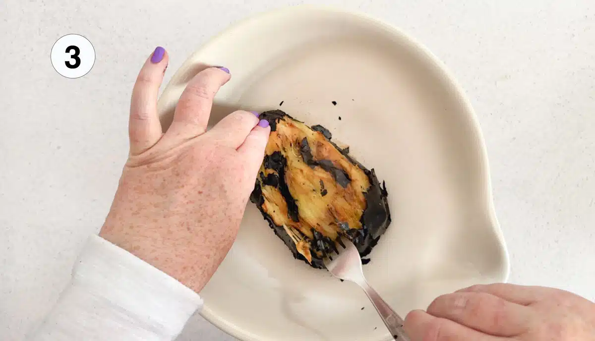 Two white hands are peeling the charred skin from a roasted eggplant that's sitting in a bowl.
