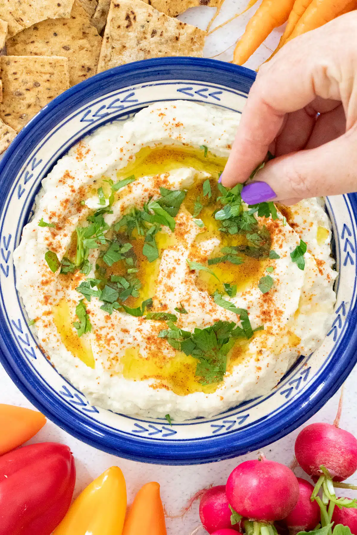 A white hand is sprinkling chopped parsley over a dish of baba ganoush. It's surrounded by vegetables and crackers.