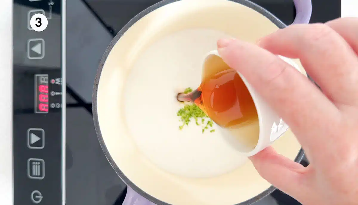 A white hand is pouring maple syrup into a small pot sitting on a stove that's filled with coconut milk and lime zest.