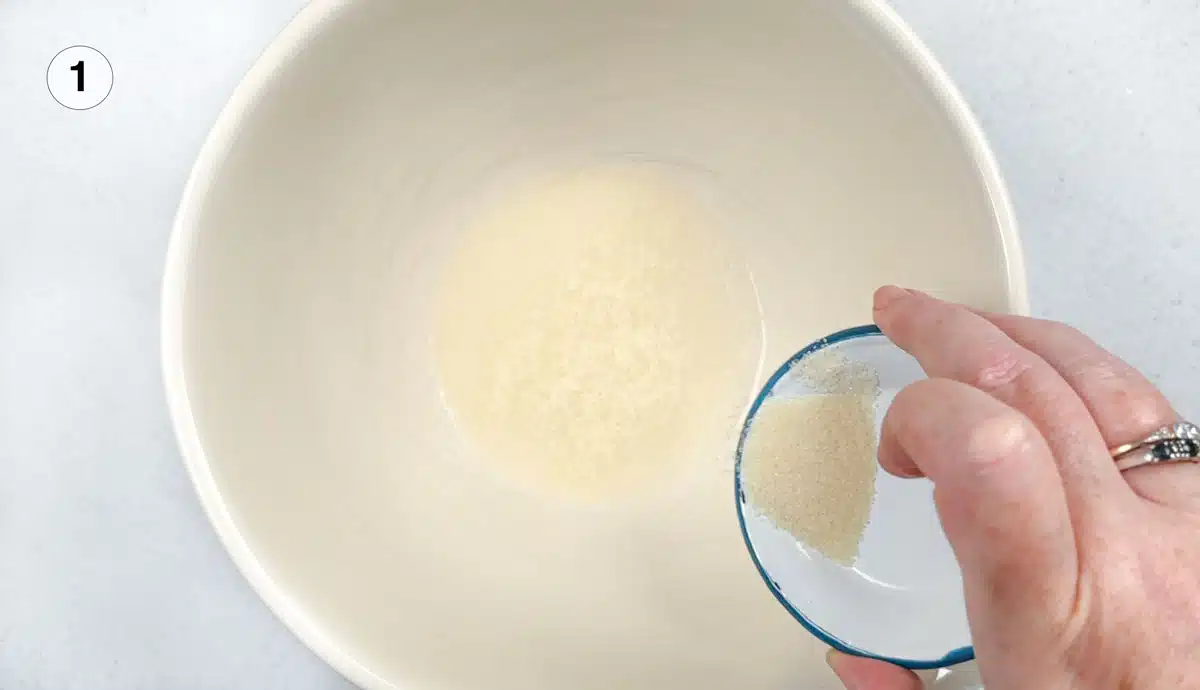 A white hand is sprinkling gelatin into a large bowl with a small amount of water in it.