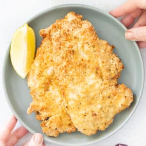 Two chicken cutlets are stacked on a grey plate. A lemon slice sits beside them.