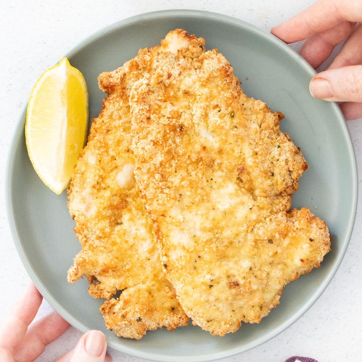 Two chicken cutlets are stacked on a grey plate. A lemon slice sits beside them.