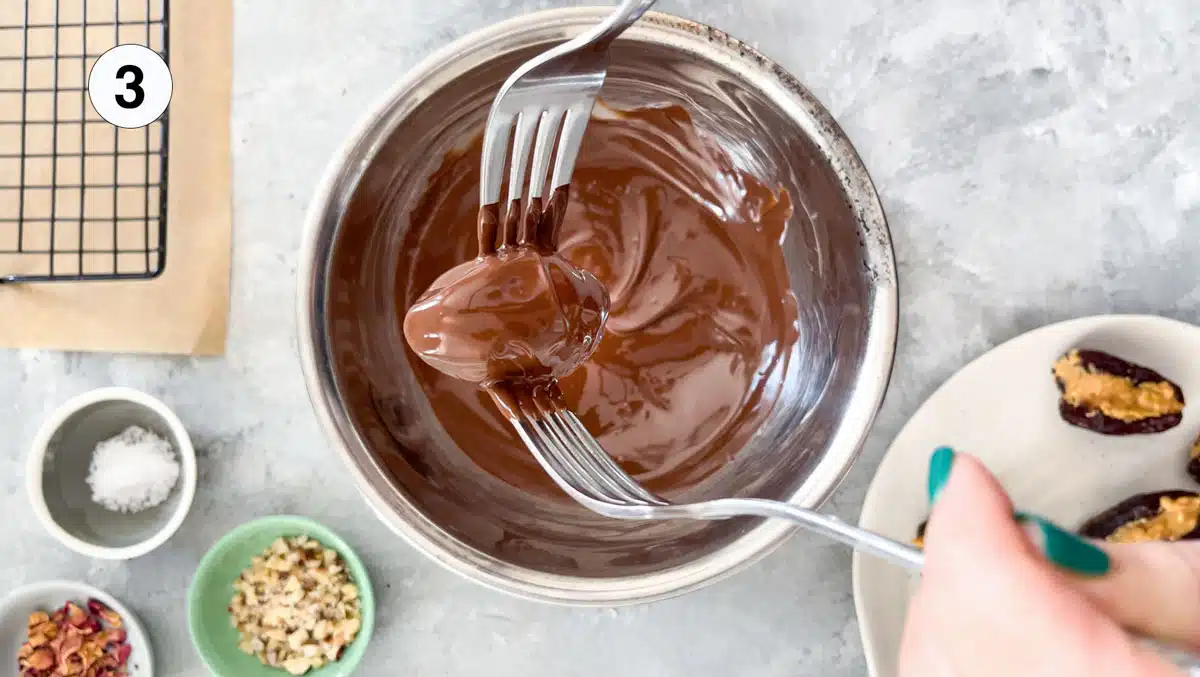 Two white hands are holding one stuffed date with two forks and dipping it in a bowl full of melted chocolate.