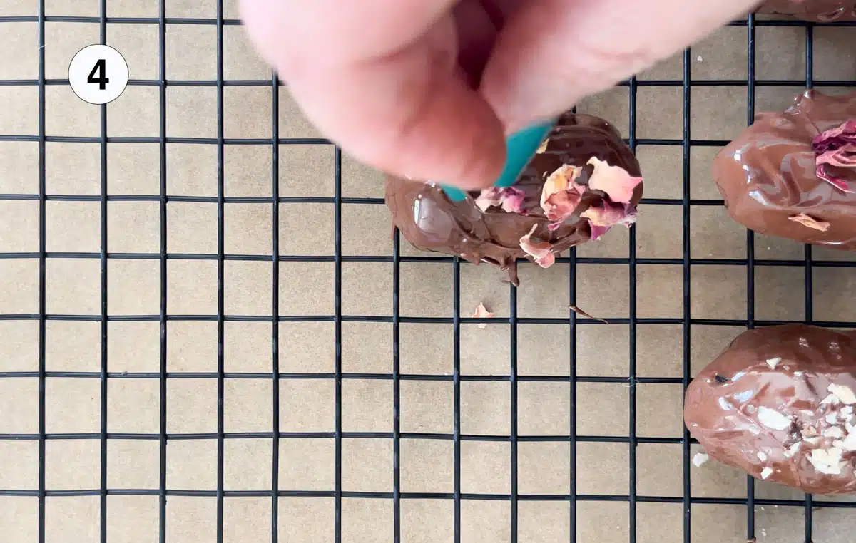 Chocolate covered dates have been placed on a wire rack and a white hand is sprinkling rose petals over one of them.