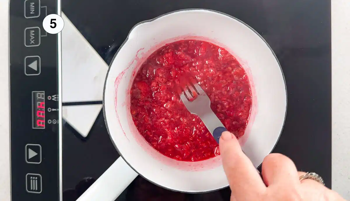 Raspberries, are being crushed under a fork in a small white saucepan on a stovetop.