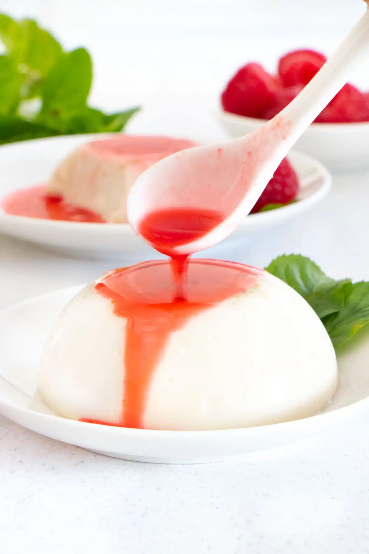 Bright red raspberry coulis is being poured over a small white coconut panna cotta sitting on a white plate.