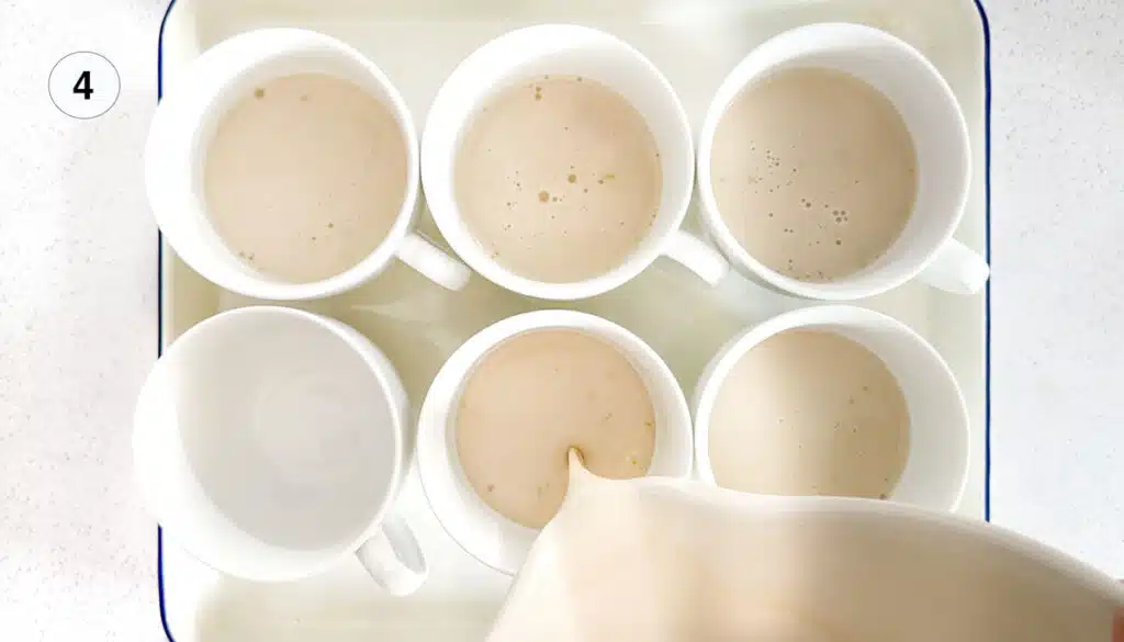 Panna cotta mix is being poured from a large bowl into six white cups sitting on a white tray.