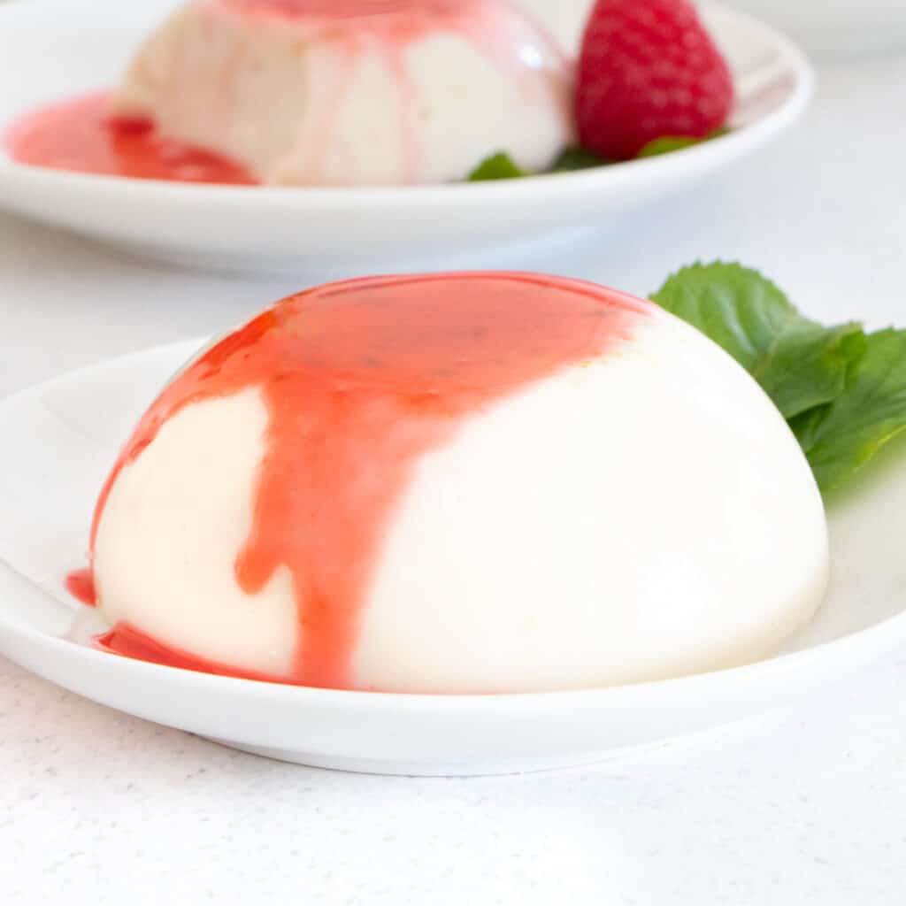 Close up of a panna cotta on a white dish that has been drizzled with raspberry sauce.