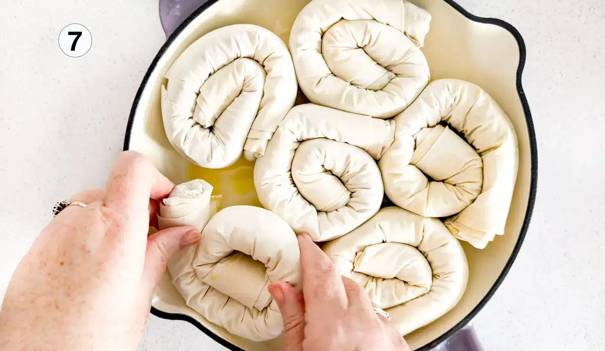 Two white hands are adding filo spirals filled with leek and feta mixture to a large skillet.