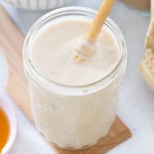Close-up of a small jar of white, whipped honey sitting on a wooden board. A wooden honey server is dipped into the jar.