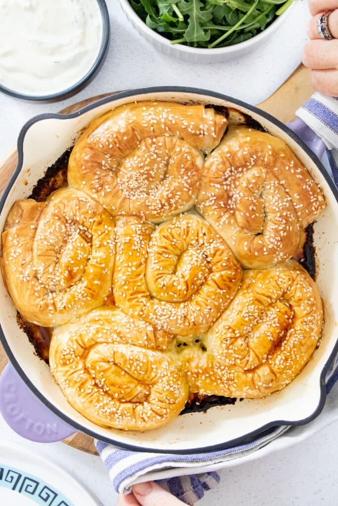 A skillet held by two hands is filled with six golden brown scrolls of Greek Leek Pie topped with sesame seeds.