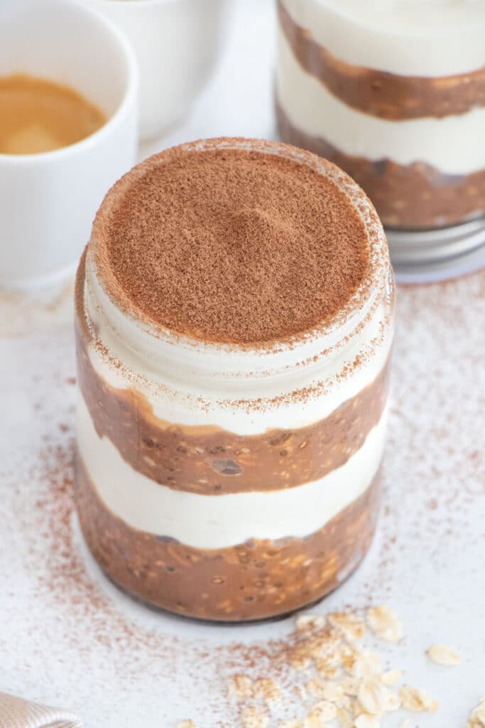 A jar of Tiramisu Overnight Oats sits on a table filled with layers of coffee chocolate oats and sweetened yoghurt.