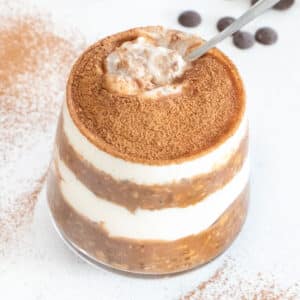 A glass of overnight oats with layers of yoghurt has just been sprinkled with cacao over the top and a spoon dipped in.