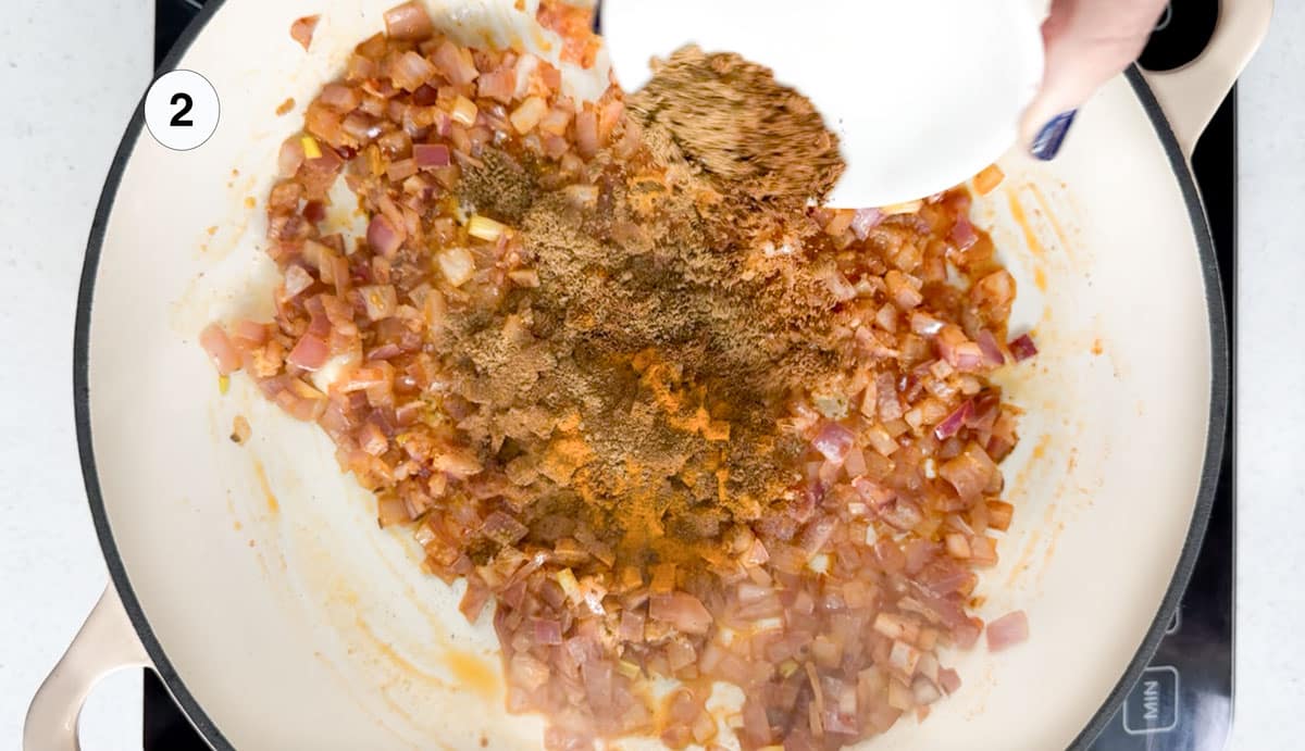 Curry spices are being added to onions and harissa simmering in a large pan.