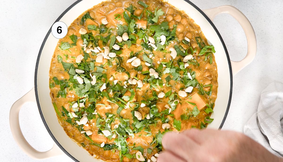 A white hand is sprinkling chopped cilantro and cashews over the top of a spinach and chickpea curry.
