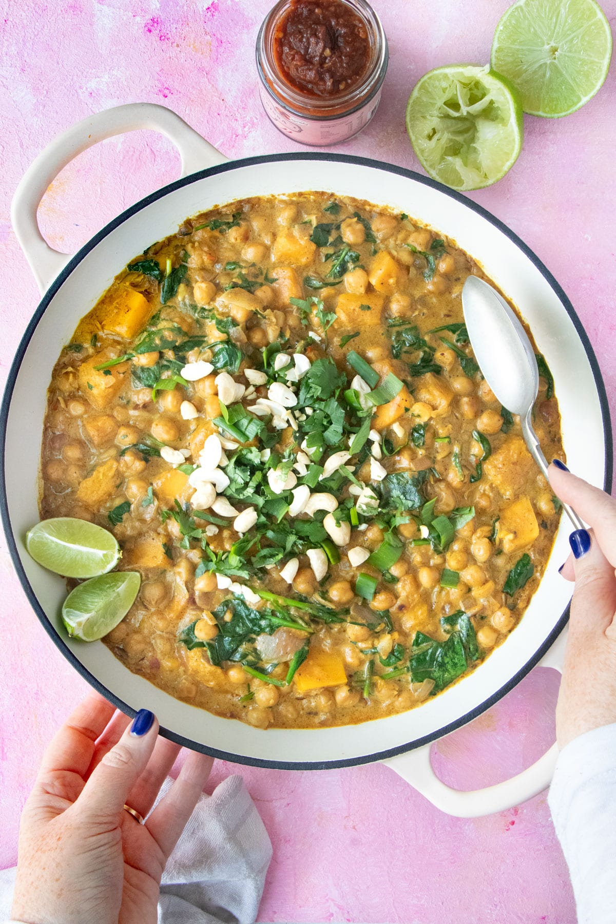 A white hand is putting a spoon into a large pot of chickpea spinach curry topped with cilantro and cashews.