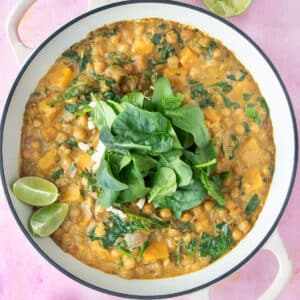 A large pot of chickpea and spinach curry is sitting on a pink tabletop. Extra spinach and lime wedges are decorating the top.